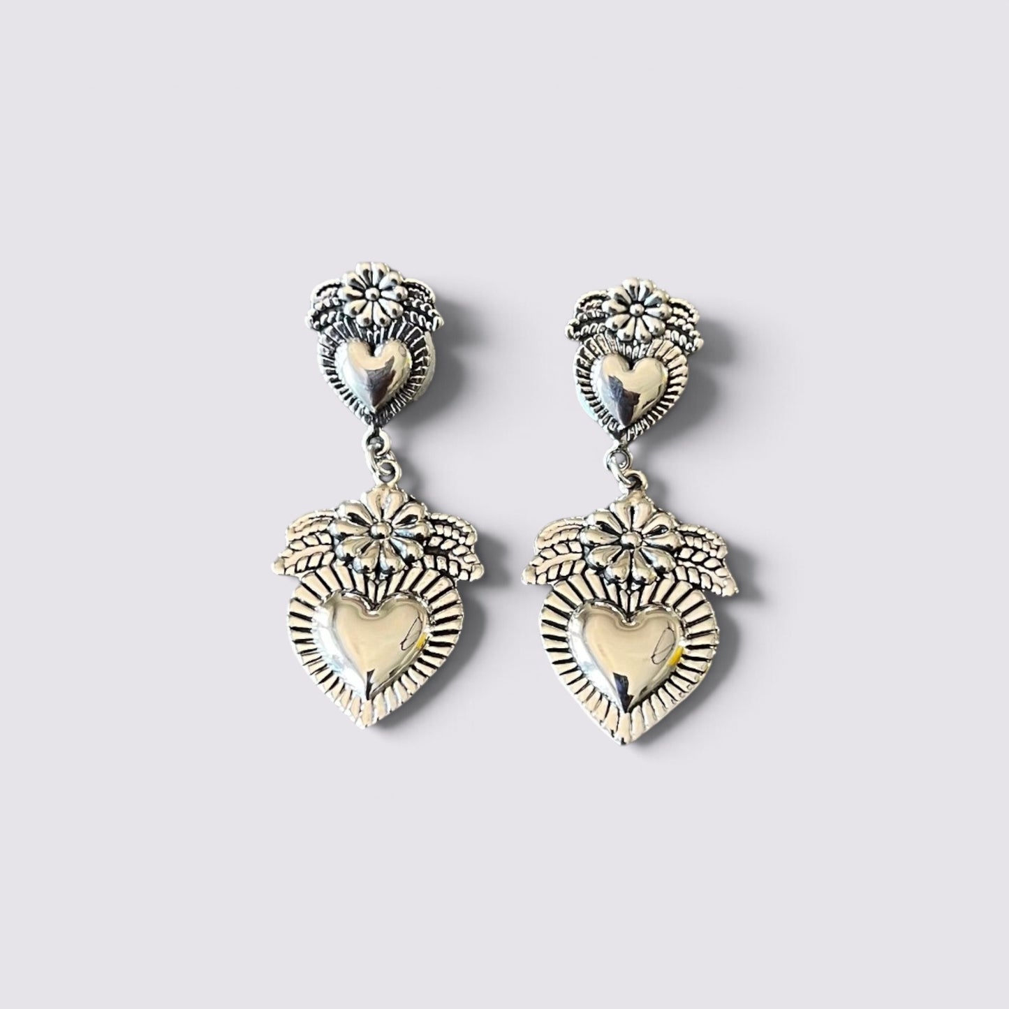 A pair of silver-toned dangle earrings featuring a heart-shaped motif with floral and sunburst details, displayed on a light gray background.