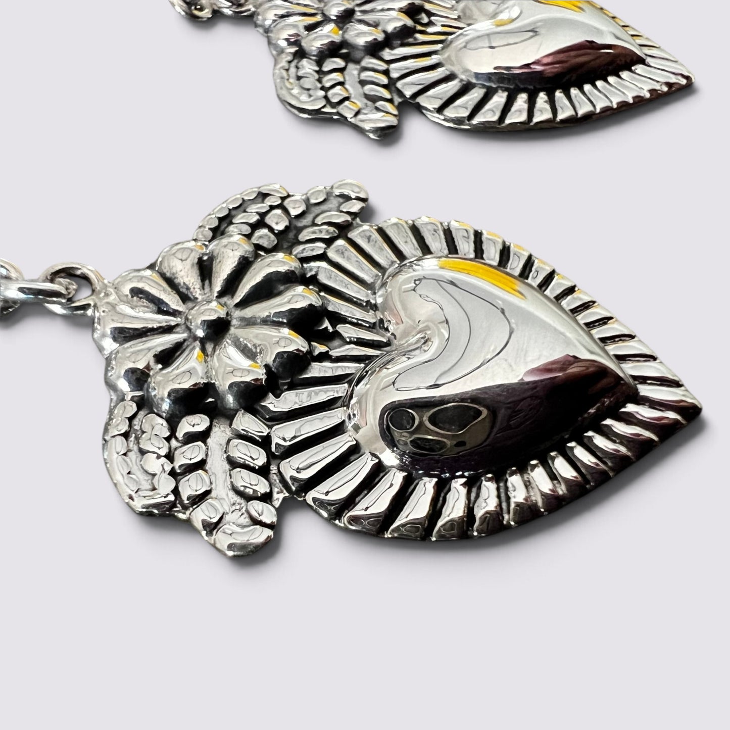 A close-up of a silver-toned metal pendant featuring a stylized heart and flower design with engraved details, shown on a light gray background.
