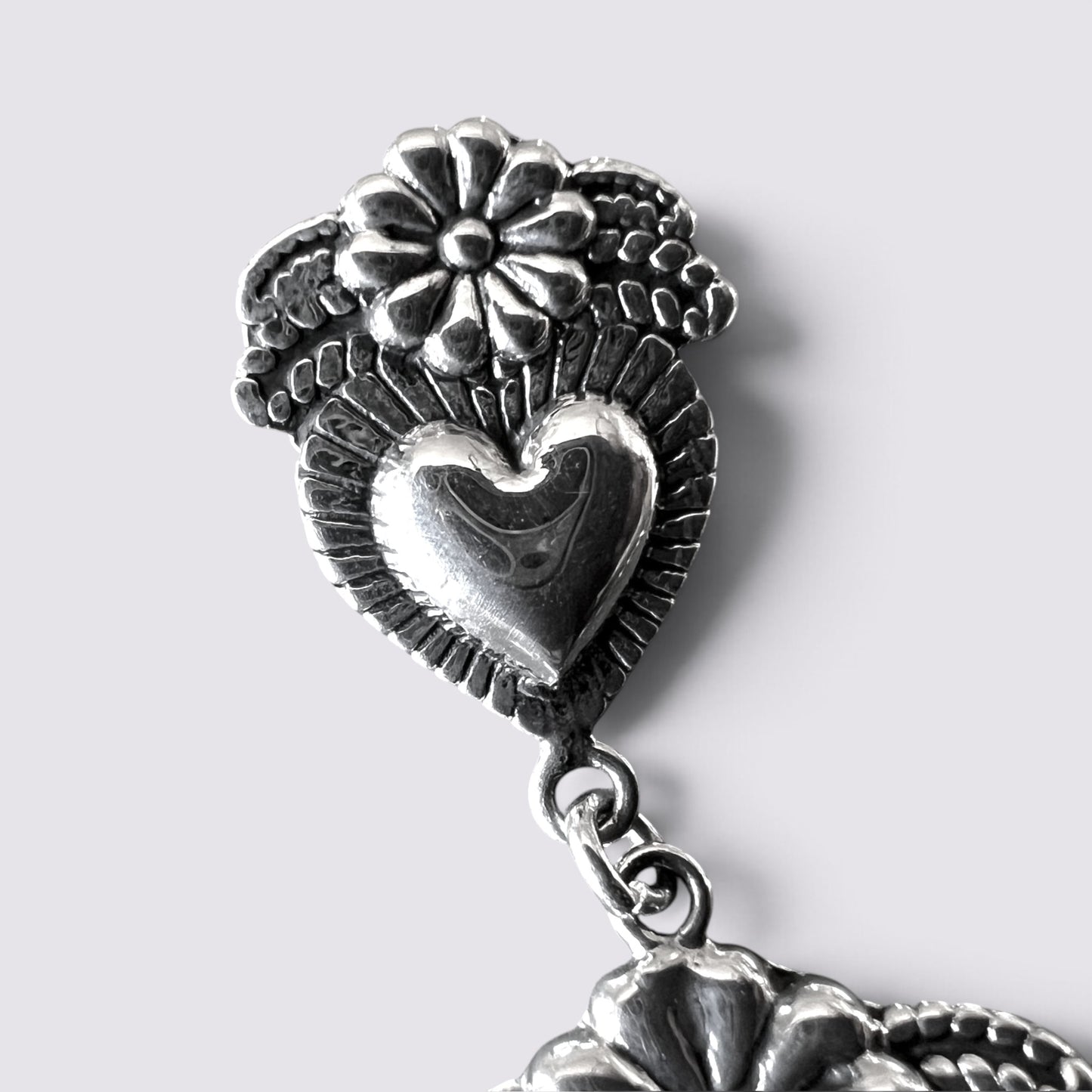 Close-up of a silver-toned metal earring featuring a heart-shaped centerpiece, textured leaf-like detailing, and a flower motif at the top, shown against a light gray background.