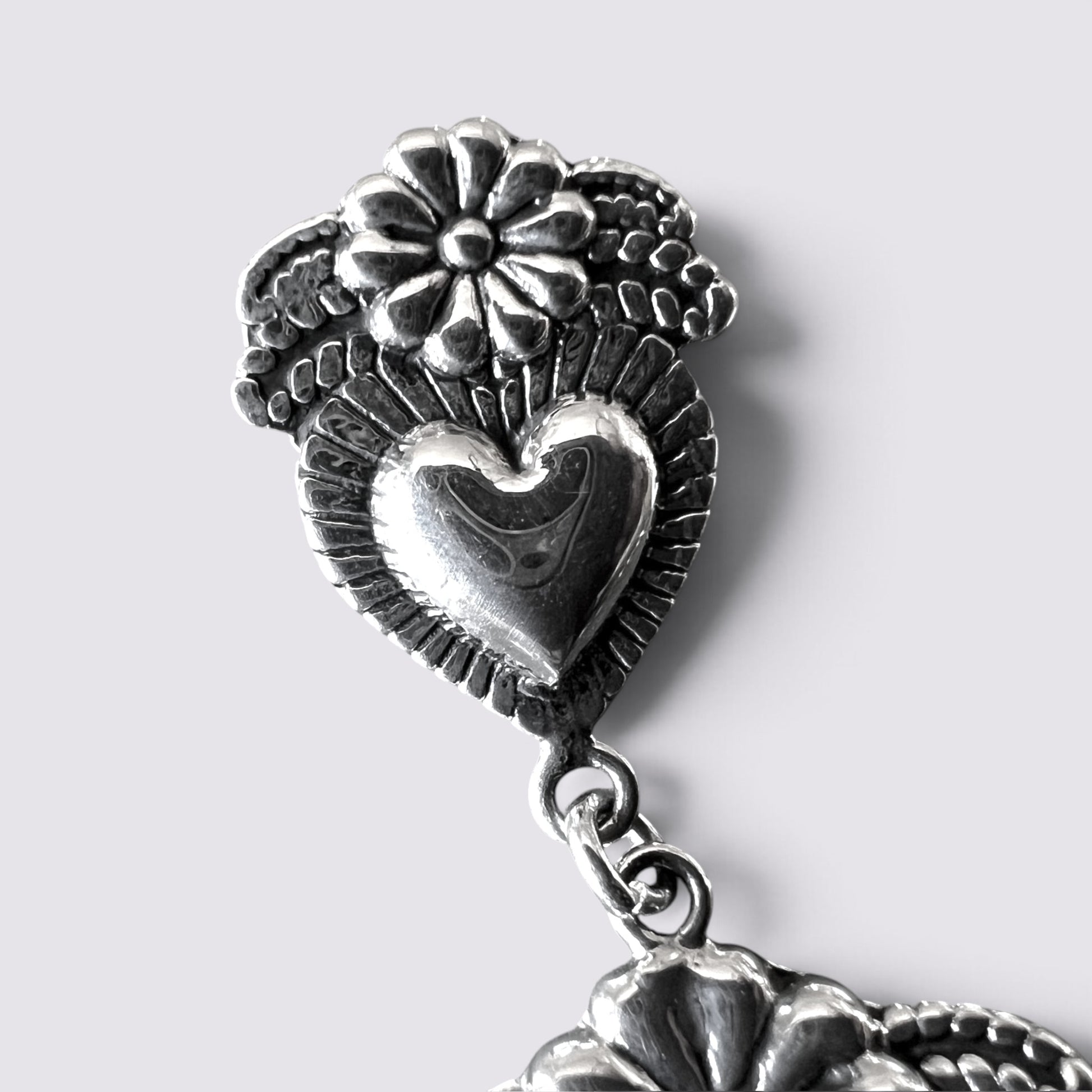 Close-up of a silver-toned metal earring featuring a heart-shaped centerpiece, textured leaf-like detailing, and a flower motif at the top, shown against a light gray background.